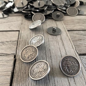 May include: A collection of silver-colored metal buttons with a vintage design. The buttons feature an embossed image of a knight on horseback, with the text "S. GEORGIUS PATRONVS" around the edge. Some buttons are scattered on a weathered wooden surface.
