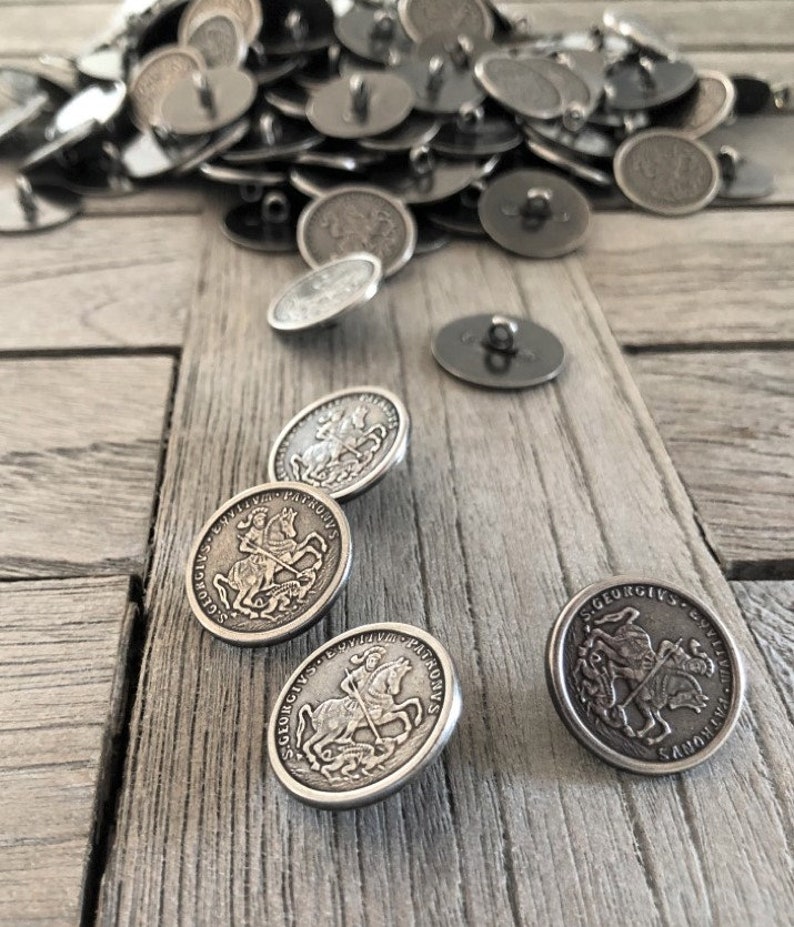 May include: A collection of silver-colored metal buttons with a vintage design. The buttons feature an embossed image of a knight on horseback, with the text "S. GEORGIUS PATRONVS" around the edge. Some buttons are scattered on a weathered wooden surface.