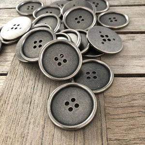 Può includere: Un mucchio di bottoni in metallo argentato con quattro fori ciascuno. I bottoni sono rotondi e hanno un bordo rialzato.