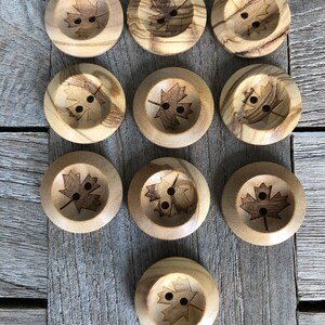 May include: Twelve wooden buttons with a maple leaf design. The buttons are a light brown color and have two holes each.