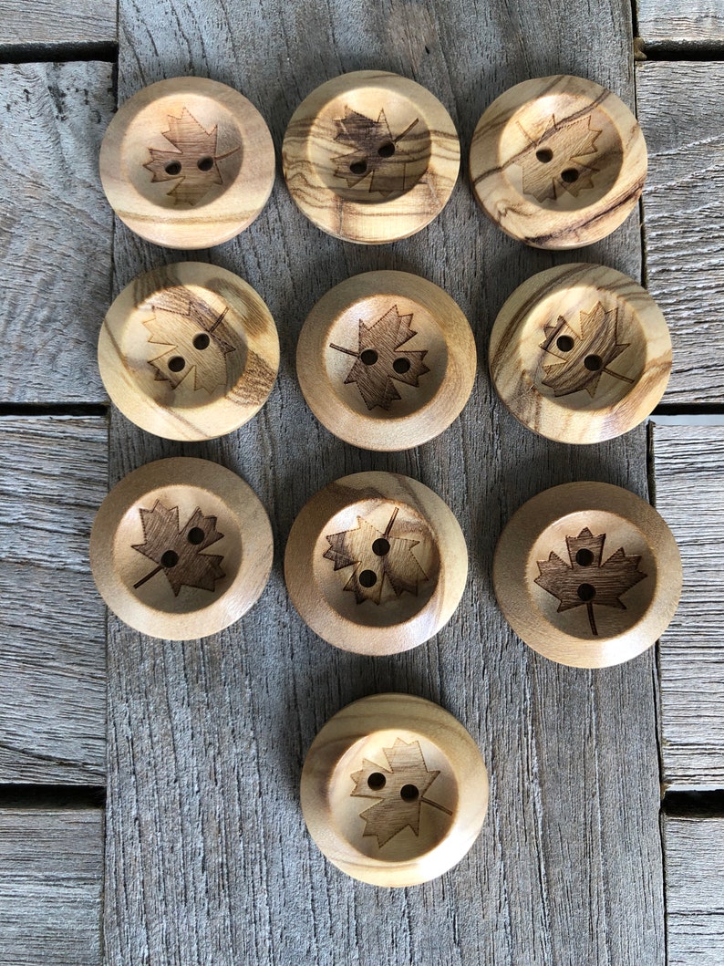 May include: Twelve wooden buttons with a maple leaf design. The buttons are a light brown color and have two holes each.