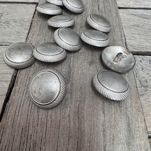 Puede incluir: Un grupo de botones de metal plateado con un diseño texturizado. Los botones están dispuestos sobre una superficie de madera.