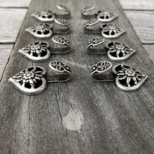 May include: A set of ten silver-toned metal hooks in the shape of a heart with a stylized flower design. The hooks are arranged in two rows of five on a wooden surface.