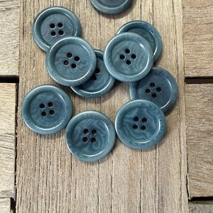 Op de afbeelding: Een verzameling van tien blauwgrijze knopen met elk vier gaatjes, geplaatst op een houten ondergrond. De knopen hebben een subtiel gemarmerd effect. Ideaal voor naai- en knutselprojecten.