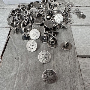Op de afbeelding: Een stapel zilverkleurige metalen knopen met een decoratief ontwerp. De knopen hebben een lus aan de achterkant om te naaien.
