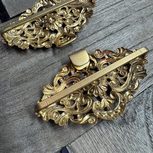May include: Two ornate, gold-colored metal hair barrettes. Each barrette features a floral and scroll design with a rectangular clasp. The barrettes are displayed on a wooden surface.