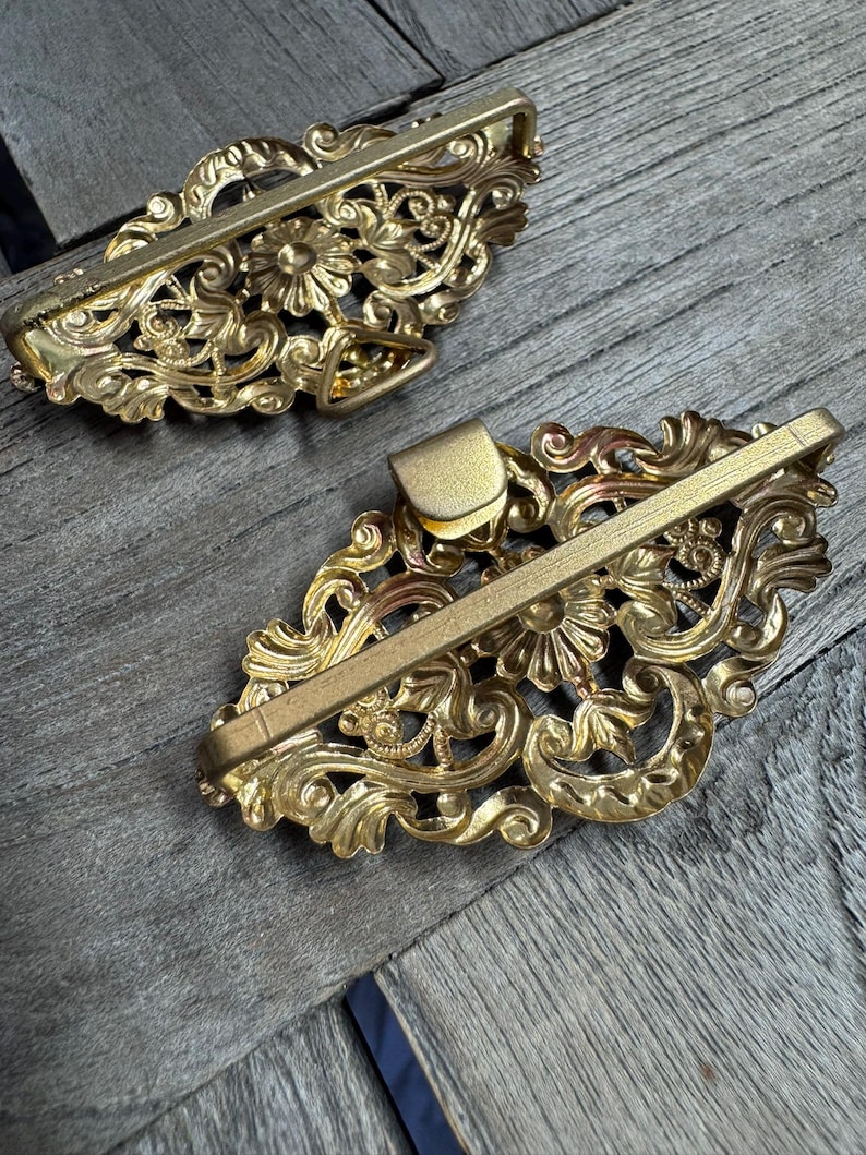 May include: Two ornate, gold-colored metal hair barrettes. Each barrette features a floral and scroll design with a rectangular clasp. The barrettes are displayed on a wooden surface.