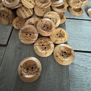 May include: A collection of wooden buttons with two holes, some featuring the engraved text "Cozy Winter" with floral accents. The buttons are light brown with a wood grain pattern, ideal for sewing and craft projects.