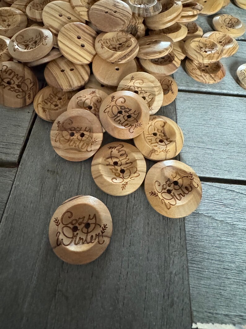 May include: A collection of wooden buttons with two holes, some featuring the engraved text "Cozy Winter" with floral accents. The buttons are light brown with a wood grain pattern, ideal for sewing and craft projects.