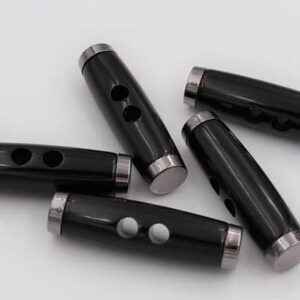 May include: A collection of five black toggle buttons, each featuring a smooth, cylindrical body with two central holes for attachment. The ends of each button are capped with polished silver-toned metal rings, providing a contrast to the black.