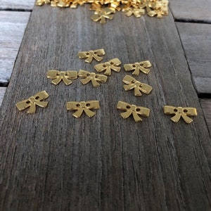 May include: A pile of gold-colored bow-shaped buttons on a wooden surface. The buttons are small and have a two-hole design.