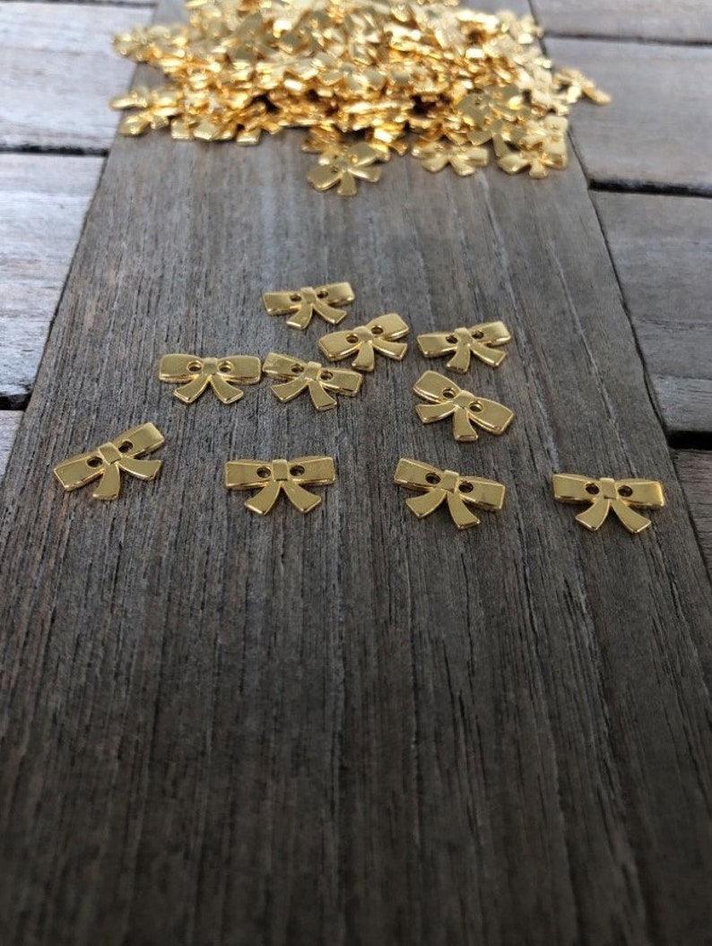 May include: A pile of gold-colored bow-shaped buttons on a wooden surface. The buttons are small and have a two-hole design.