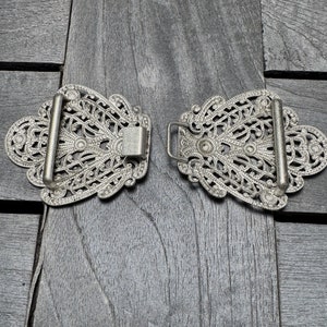 K&ouml;nnte beinhalten: Ein Paar silberne G&uuml;rtelschnallen mit filigranen Designs. Die Schnallen sind verziert und detailliert, mit einem zarten Blumenmuster.