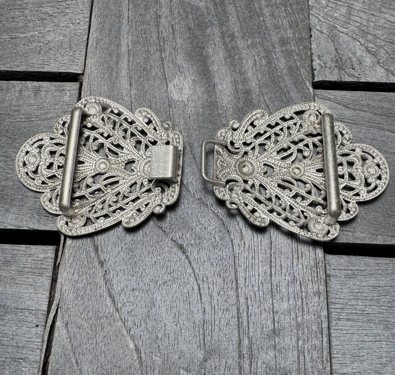 K&ouml;nnte beinhalten: Ein Paar silberne G&uuml;rtelschnallen mit filigranen Designs. Die Schnallen sind verziert und detailliert, mit einem zarten Blumenmuster.