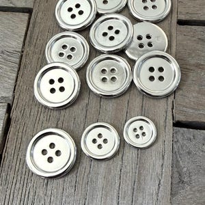 May include: A selection of shiny, silver buttons with four holes. The buttons are of varying sizes and are arranged on a weathered wooden surface. Each button has a raised rim and a smooth, reflective finish.