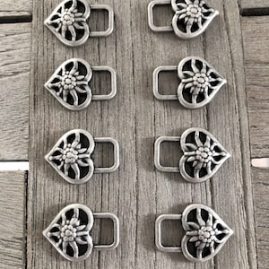May include: Eight silver-toned metal clasps. Each clasp features a heart-shaped design with a floral center and a rectangular loop. The clasps are arranged on a weathered wooden surface, showcasing their intricate details and metallic sheen.