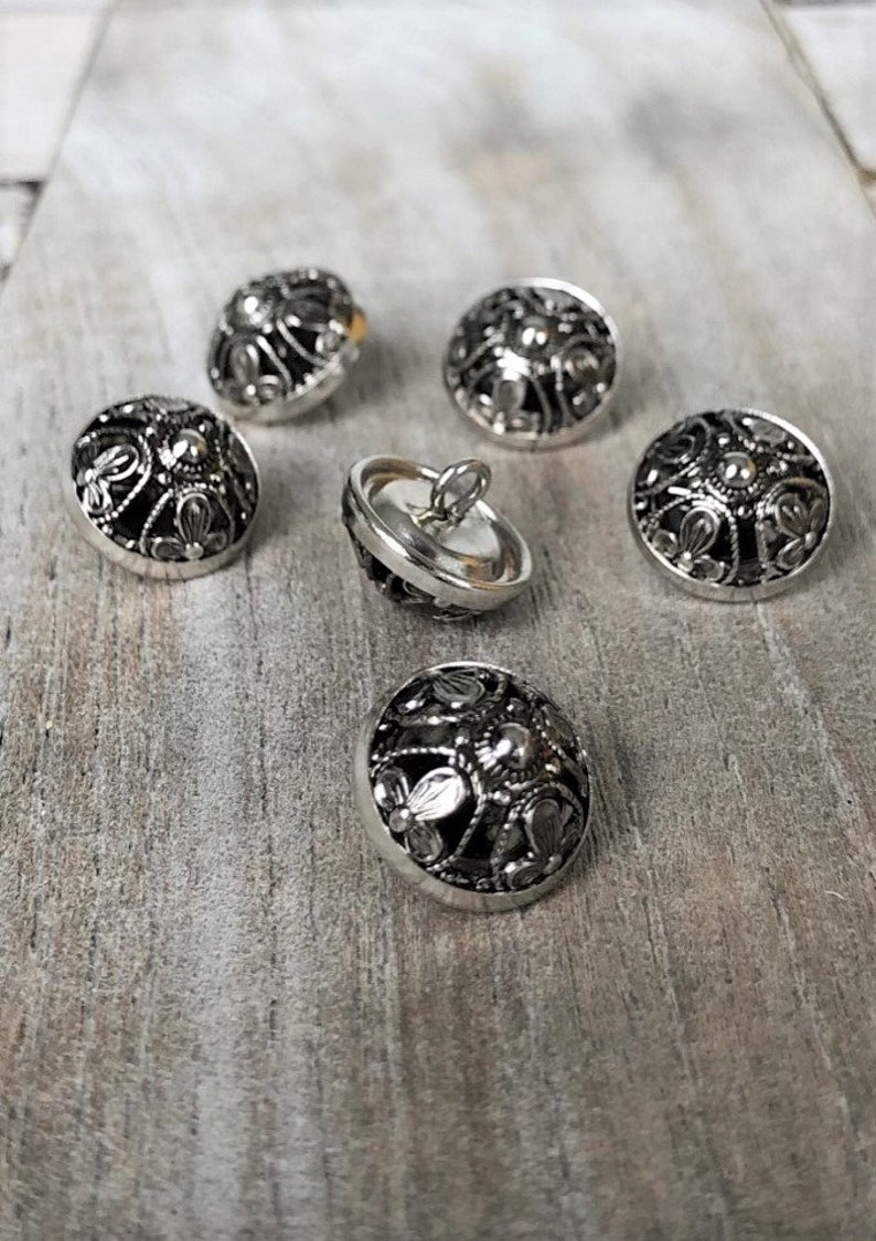 May include: Six silver-toned buttons with intricate black filigree designs. The buttons are round and have a domed shape.