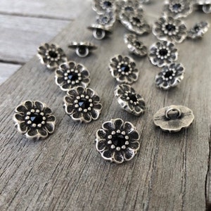 Puede incluir: Una colección de botones de metal plateados con centros de pedrería negra. Los botones tienen forma de flores con múltiples pétalos.