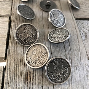 Könnte beinhalten: Eine Sammlung antiker, silberfarbener Knöpfe mit einem erhabenen Design eines Ritters zu Pferd. Die runden Knöpfe mit detailliertem Rand liegen auf einer verwitterten Holzoberfläche.