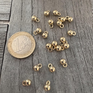 Könnte beinhalten: Eine Sammlung von 18 goldfarbenen Metallperlen mit einer Öse oben. Die Perlen sind auf einer Holzoberfläche verstreut.