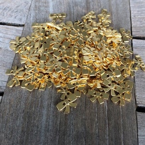 May include: A large pile of gold-colored metal bow-shaped embellishments. The bows are small and have two holes in the center.