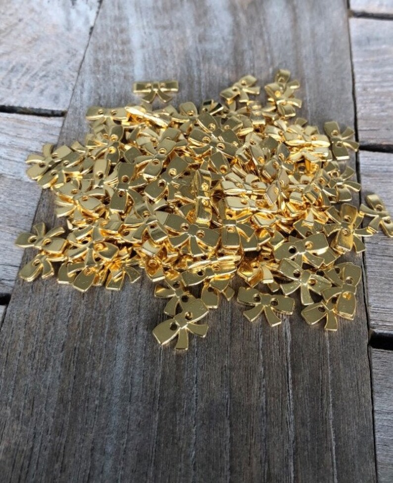 May include: A large pile of gold-colored metal bow-shaped embellishments. The bows are small and have two holes in the center.