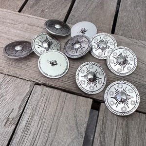 Könnte beinhalten: Eine Sammlung silberfarbener Metallknöpfe mit Sonnenstrahlendesign. Die Knöpfe haben einen zentralen, erhabenen Stift und einen dekorativen Rand. Die Knöpfe sind auf einer Holzoberfläche verstreut.