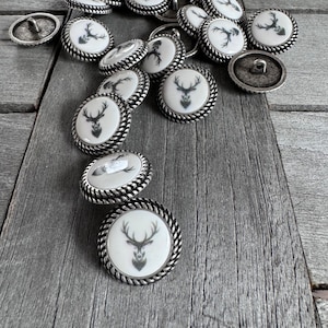 Può includere: Una collezione di bottoni argentati con sfondo bianco e un design a silhouette di cervo nero. I bottoni hanno un bordo a corda.