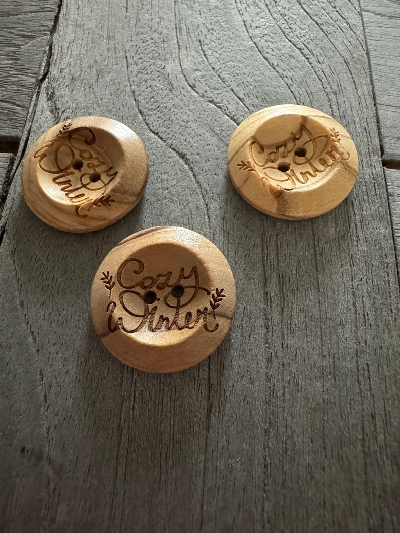 May include: Three round wooden buttons with the words "Cozy Winter" etched into the surface. The buttons are light brown with two holes for sewing. The text is surrounded by decorative leaves.