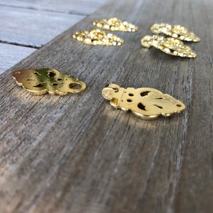 May include: A set of eight gold-toned metal decorative embellishments with intricate cut-out designs. The embellishments are shaped like a stylized flower with a loop at the top.
