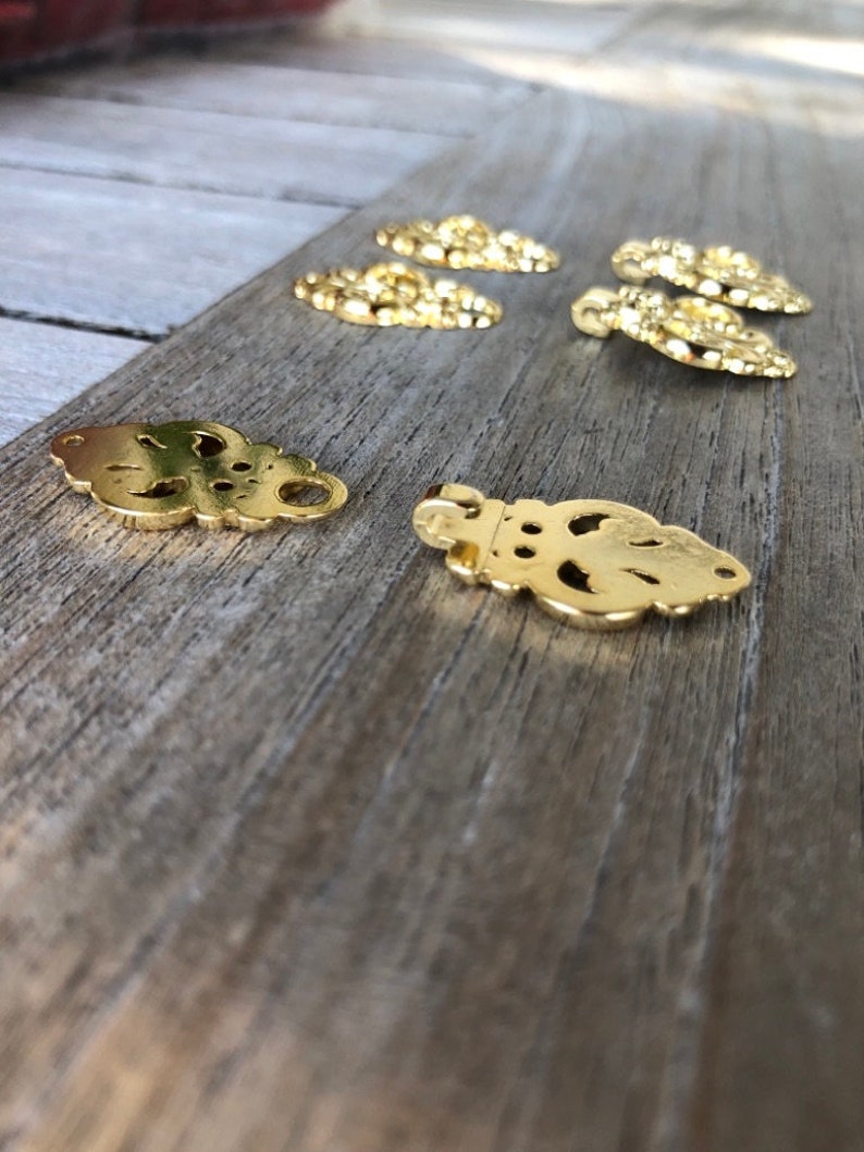 May include: A set of eight gold-toned metal decorative embellishments with intricate cut-out designs. The embellishments are shaped like a stylized flower with a loop at the top.
