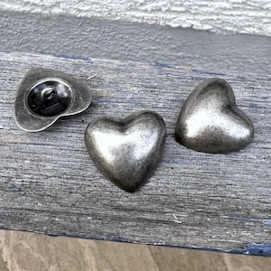 May include: Three silver heart-shaped buttons with a textured finish. The buttons are arranged on a wooden surface.