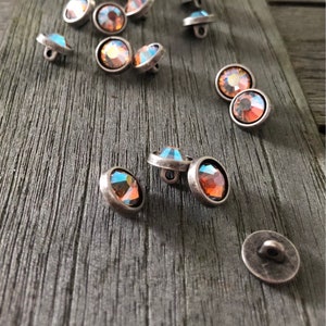 Puede incluir: Un grupo de botones redondos multifacéticos iridiscentes con ajustes plateados. Los botones están esparcidos sobre una superficie de madera.