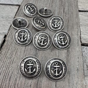Peut inclure: Dix boutons en métal argenté avec une bordure en forme de corde. Chaque bouton présente une ancre noire avec le texte "PORTS DE BRETAGNE" autour.