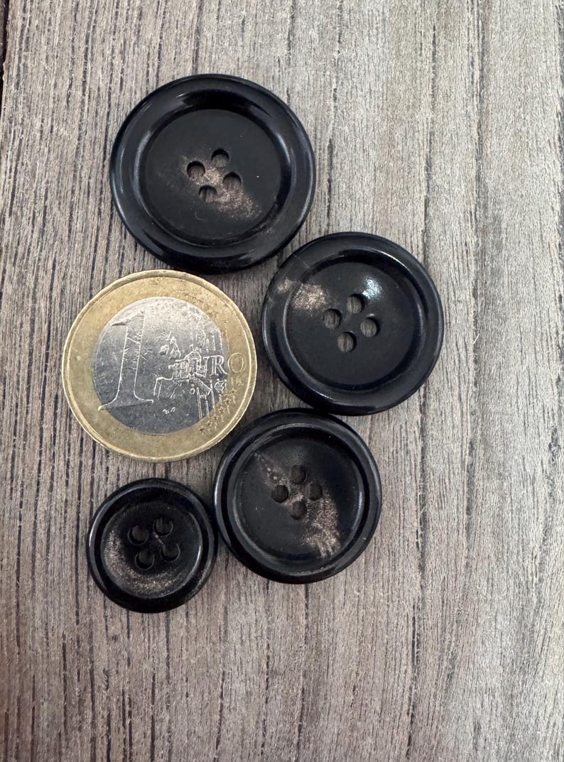 K&ouml;nnte beinhalten: Vier schwarze Kn&ouml;pfe mit einer marmorierten Oberfl&auml;che und jeweils vier L&ouml;chern. Die Kn&ouml;pfe sind in einem Halbkreis mit einer Euro-M&uuml;nze in der Mitte angeordnet.