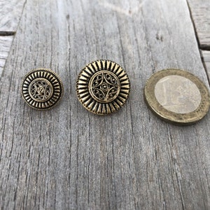 May include: Three gold-colored metal buttons with intricate designs. The buttons are round and have a raised center with a floral pattern. The buttons are arranged in a row from largest to smallest.