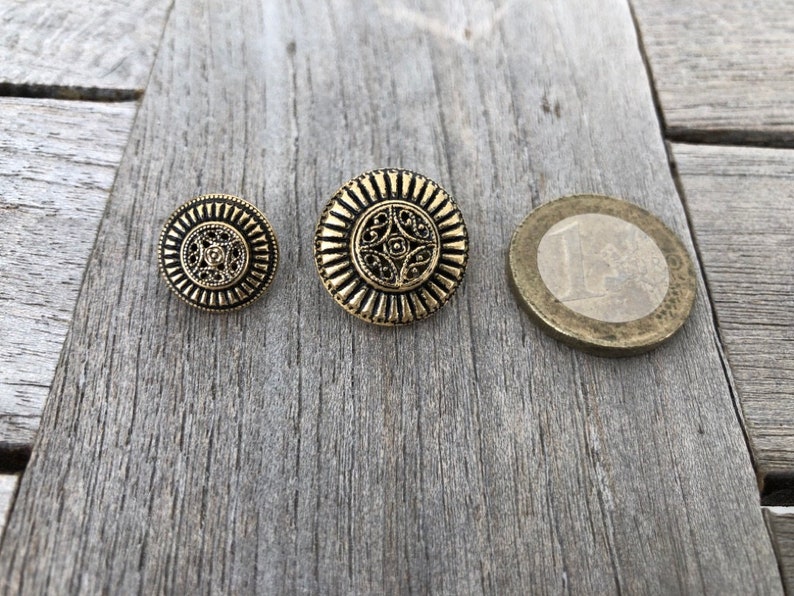 May include: Three gold-colored metal buttons with intricate designs. The buttons are round and have a raised center with a floral pattern. The buttons are arranged in a row from largest to smallest.