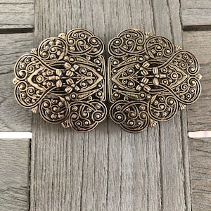 Puede incluir: Cierre metálico ornamentado de estilo antiguo en tono dorado. El cierre consta de dos mitades simétricas, cada una con intrincados diseños florales y de volutas. El cierre está diseñado para conectar y asegurar dos piezas de tela o material.