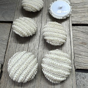 May include: A collection of six white, round buttons with a pearl and silver bead design. One button has a white backing with the number 40. The buttons are arranged on a weathered wooden surface.