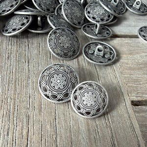 Puede incluir: Un conjunto de botones de metal plateado con un diseño floral ornamentado. Los botones redondos tienen un lazo en la parte posterior para coser. Los botones, de diferentes tamaños, están sobre una superficie de madera.