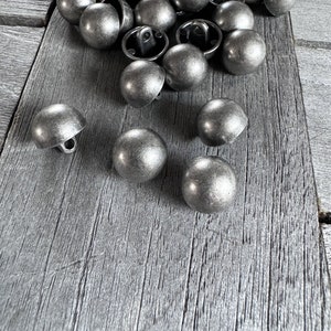 May include: A collection of antique silver-coloured dome buttons with a loop on the back. The buttons are scattered on a weathered grey wooden surface. The buttons have a rounded, textured appearance.