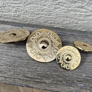 Könnte beinhalten: Vier goldfarbene, dekorative Metallscheiben mit filigranen Designs. Die Scheiben haben eine erhabene Mitte und ein kreisförmiges Muster am Rand.