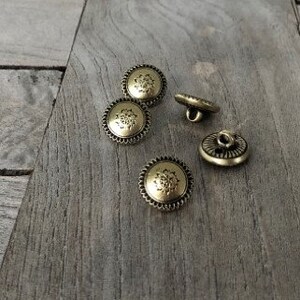 May include: Four gold-toned buttons with a decorative floral design. The buttons have a raised edge and a flat back.