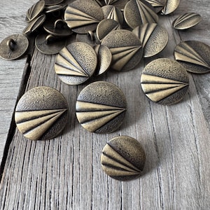 Peut inclure: Une collection de boutons antiques de couleur laiton. Les boutons sont ronds avec une surface texturée et un motif en forme d'éventail. Certains boutons ont une boucle au dos pour la couture. Les boutons sont dispersés sur une surface en bois.