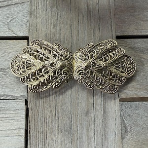 May include: A gold-colored, ornate clasp with a filigree design. The clasp is made of two symmetrical halves that connect in the center. The intricate details include swirling patterns, dots, and floral elements. The clasp is likely for a garment or accessory.