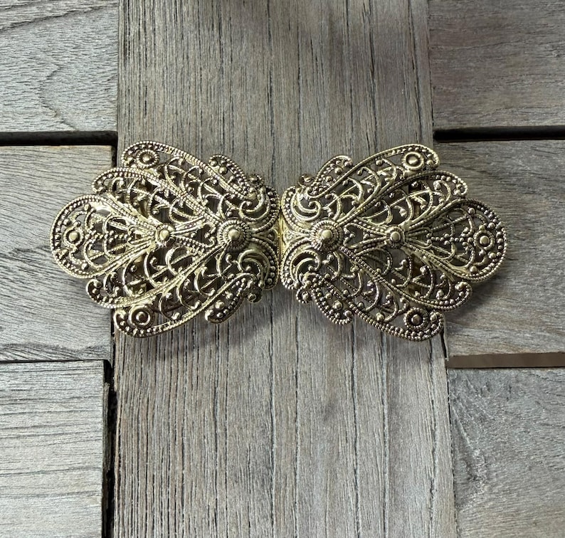 May include: A gold-colored, ornate clasp with a filigree design. The clasp is made of two symmetrical halves that connect in the center. The intricate details include swirling patterns, dots, and floral elements. The clasp is likely for a garment or accessory.