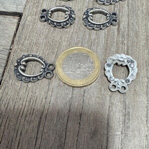 May include: Five silver-toned metal clasps with ornate designs. The clasps are shaped like a horseshoe with two loops on the top. A euro coin is included for size reference.
