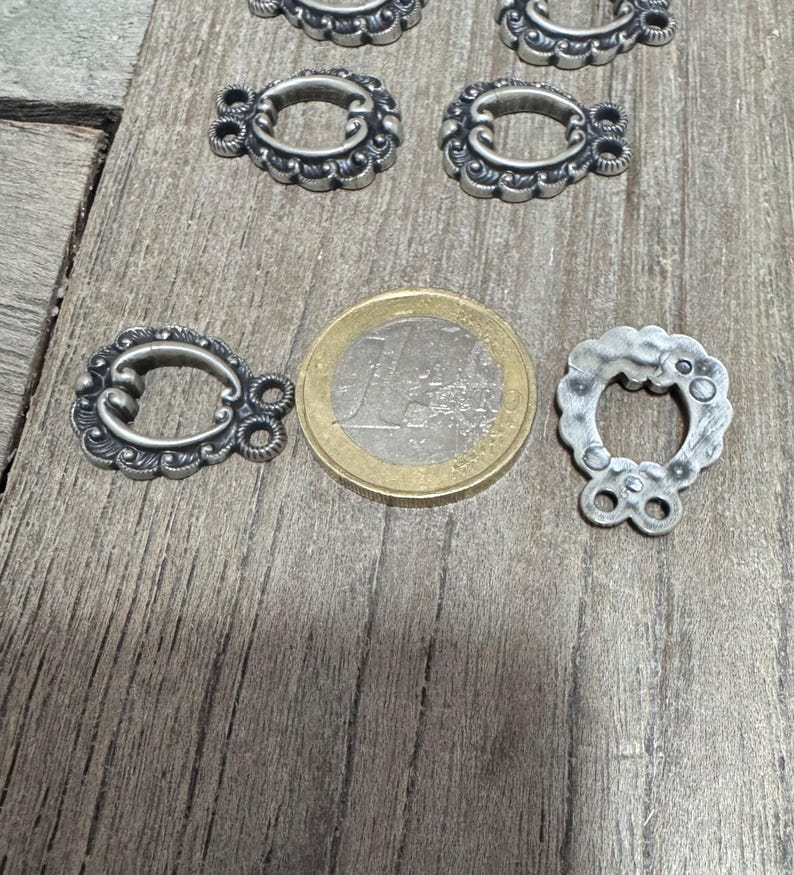 May include: Five silver-toned metal clasps with ornate designs. The clasps are shaped like a horseshoe with two loops on the top. A euro coin is included for size reference.