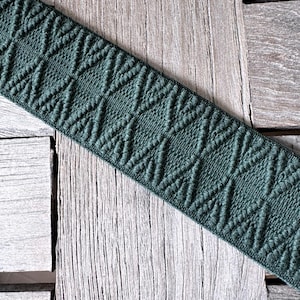 Könnte beinhalten: Ein Stück dunkelgrüner elastischer Besatz mit einem sich wiederholenden Rautenmuster. Der Besatz ist diagonal auf einer verwitterten Holzoberfläche angeordnet, wodurch das strukturierte Design und die Farbe hervorgehoben werden. Geeignet für Näh- und Bastelprojekte.