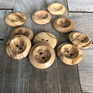 May include: A collection of ten wooden buttons with a maple leaf design. The buttons are a light brown color with darker brown wood grain patterns.
